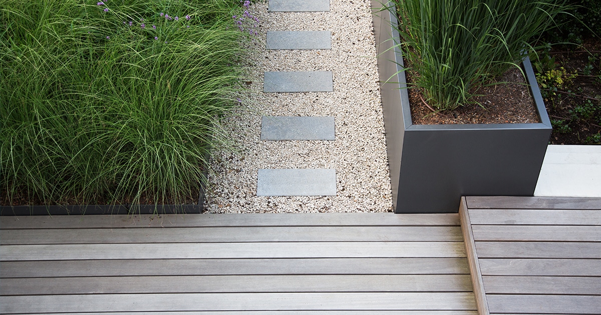Abords finis d'une maison, terrasse en bois, bande de gravier drainante et bacs métalliques alignés sur le même niveau